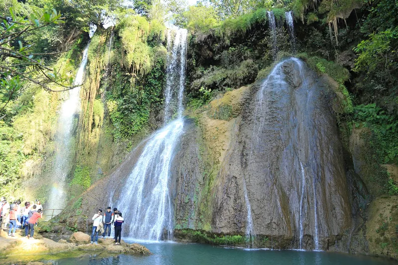 Visitors gather beneath multiple cascading waterfalls flowing over a moss-covered cliff into a turquoise pool in Moc Chau.