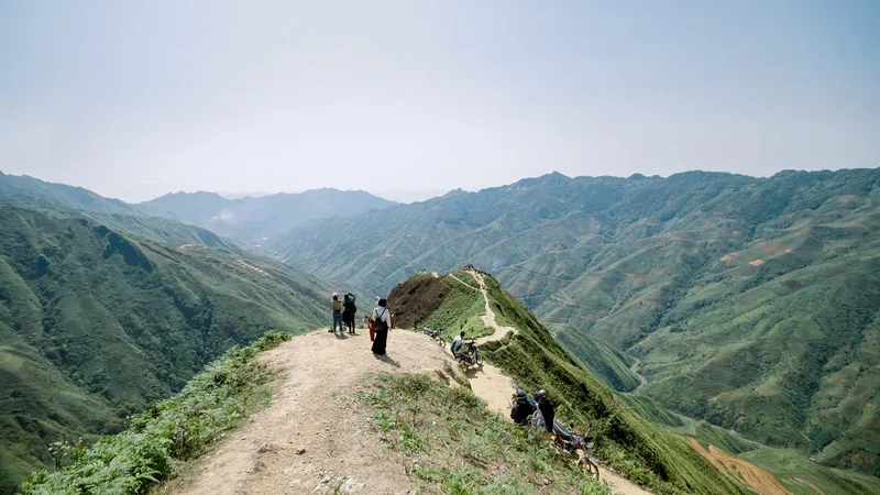 Travelers walk a narrow ridge trail with motorbikes overlooking layered green mountains at Ta Xua, Son La.